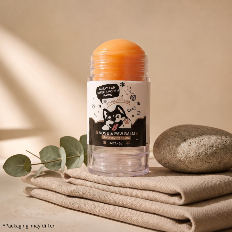 A clear plastic jar of dog nose and paw balm with an orange balm stick, set on a beige linen cloth with eucalyptus leaves and a stone against a warm beige background.