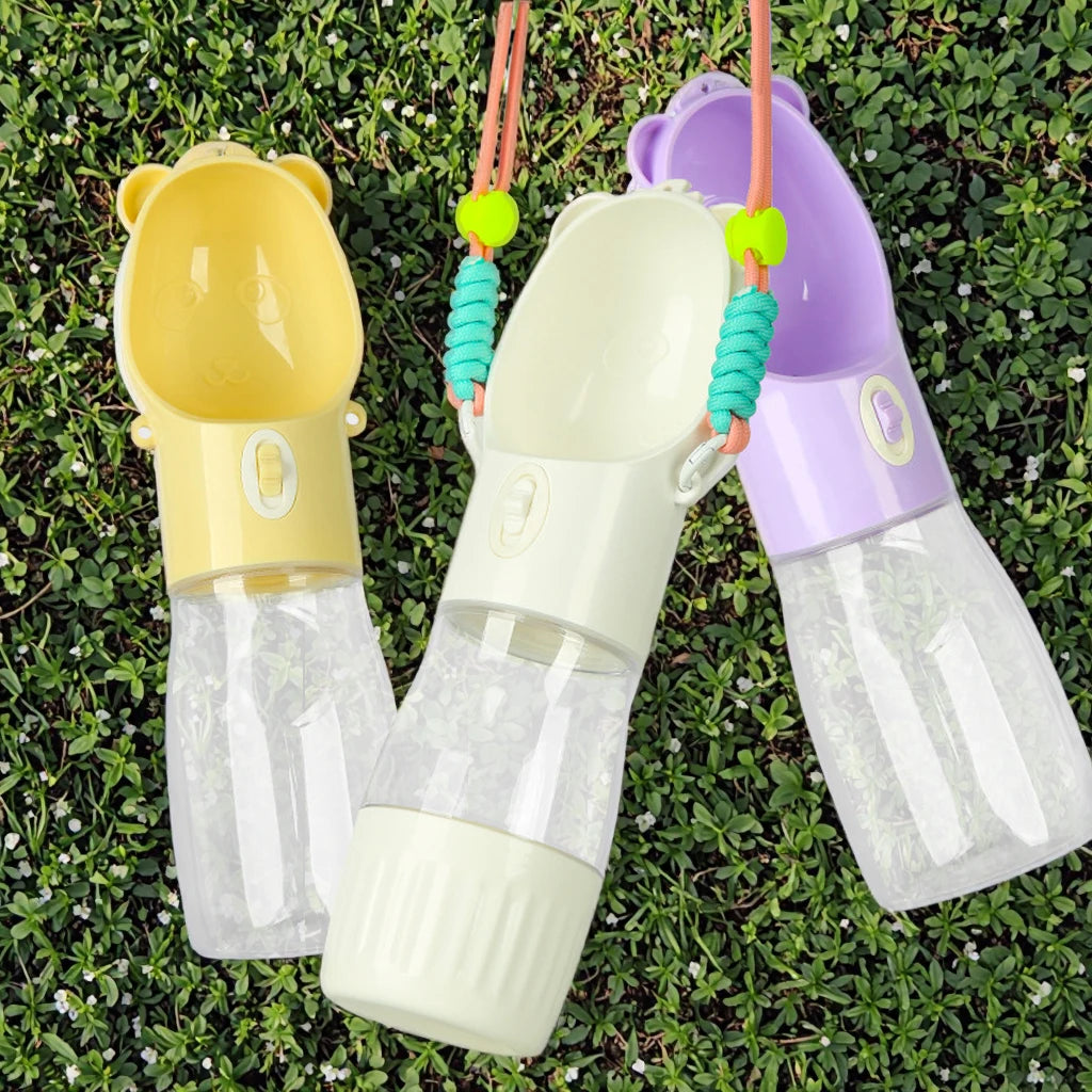 Three pet water bottles with bear-shaped caps in yellow, cream, and purple, laid out on green grass. Each bottle has a clear water reservoir, a colored upper body, and a turquoise beaded strap.