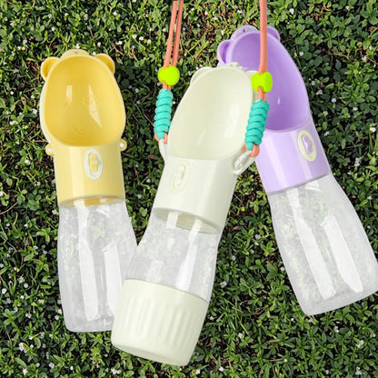 Three pet water bottles with bear-shaped caps in yellow, cream, and purple, laid out on green grass. Each bottle has a clear water reservoir, a colored upper body, and a turquoise beaded strap.