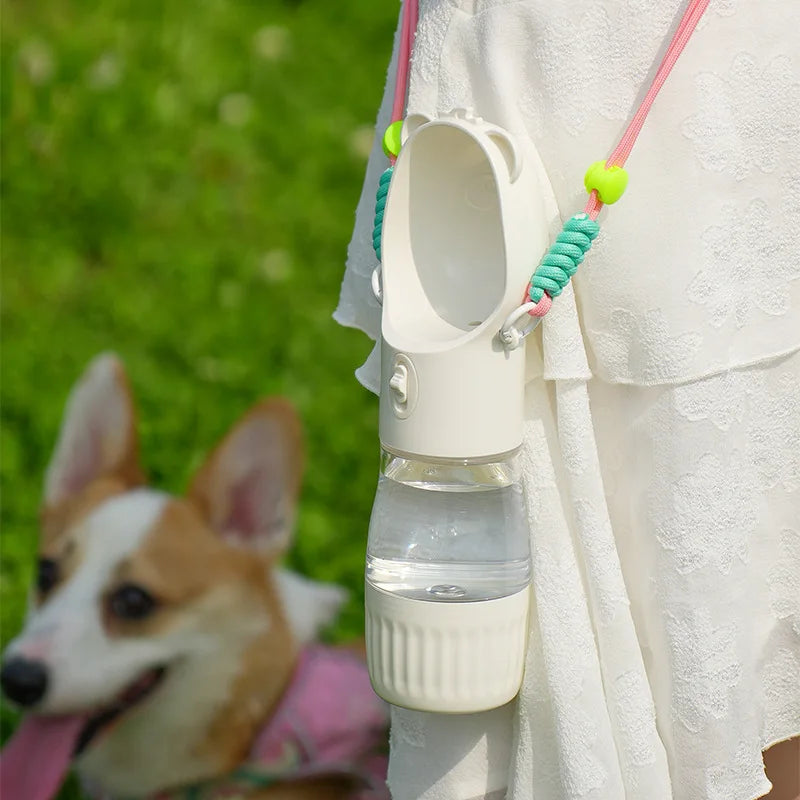 A pet water bottle holder and water bottle worn around a person's waist, with a small dog blurred in the background on grass.